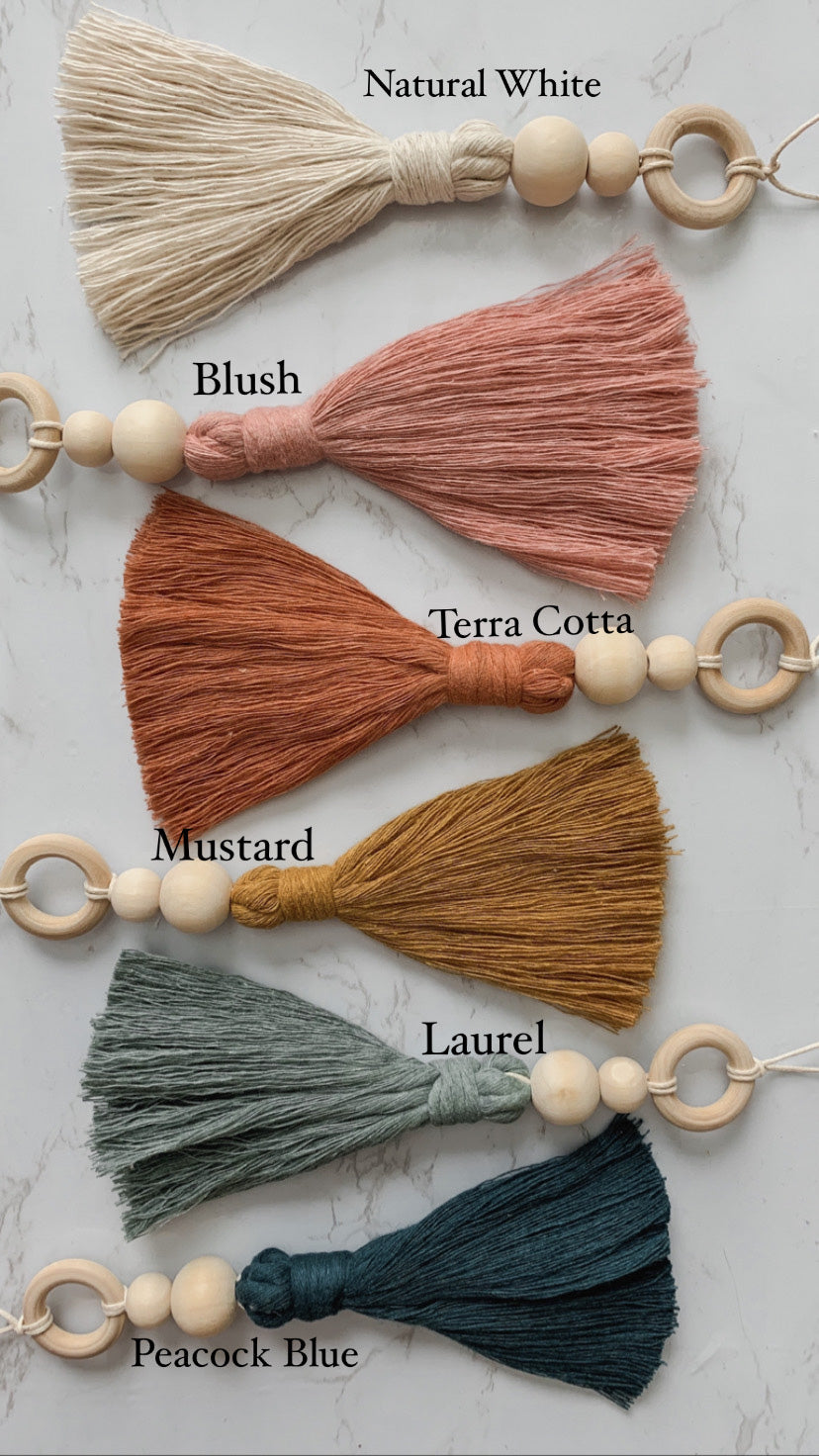 Six hanging tassel charms with wooden beading detail shown in white, blush, terra cotta, mustard, teal, and blue colors 