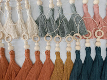 Load image into Gallery viewer, Twelve hanging tassel charms with wooden beading detail shown in white, blush, terra cotta, mustard, teal, and blue colors 
