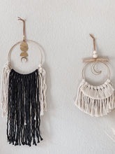 Load image into Gallery viewer, A macrame wall hanging with natural white and black colored fringe hand knotted on a gold ring with brass charms perfectly placed next to a smaller macrame wall hanging

