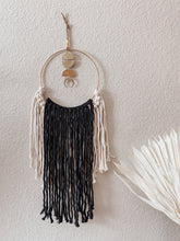 Load image into Gallery viewer, A macrame wall hanging with natural white and black colored fringe hand knotted on a gold ring with brass charms perfectly placed

