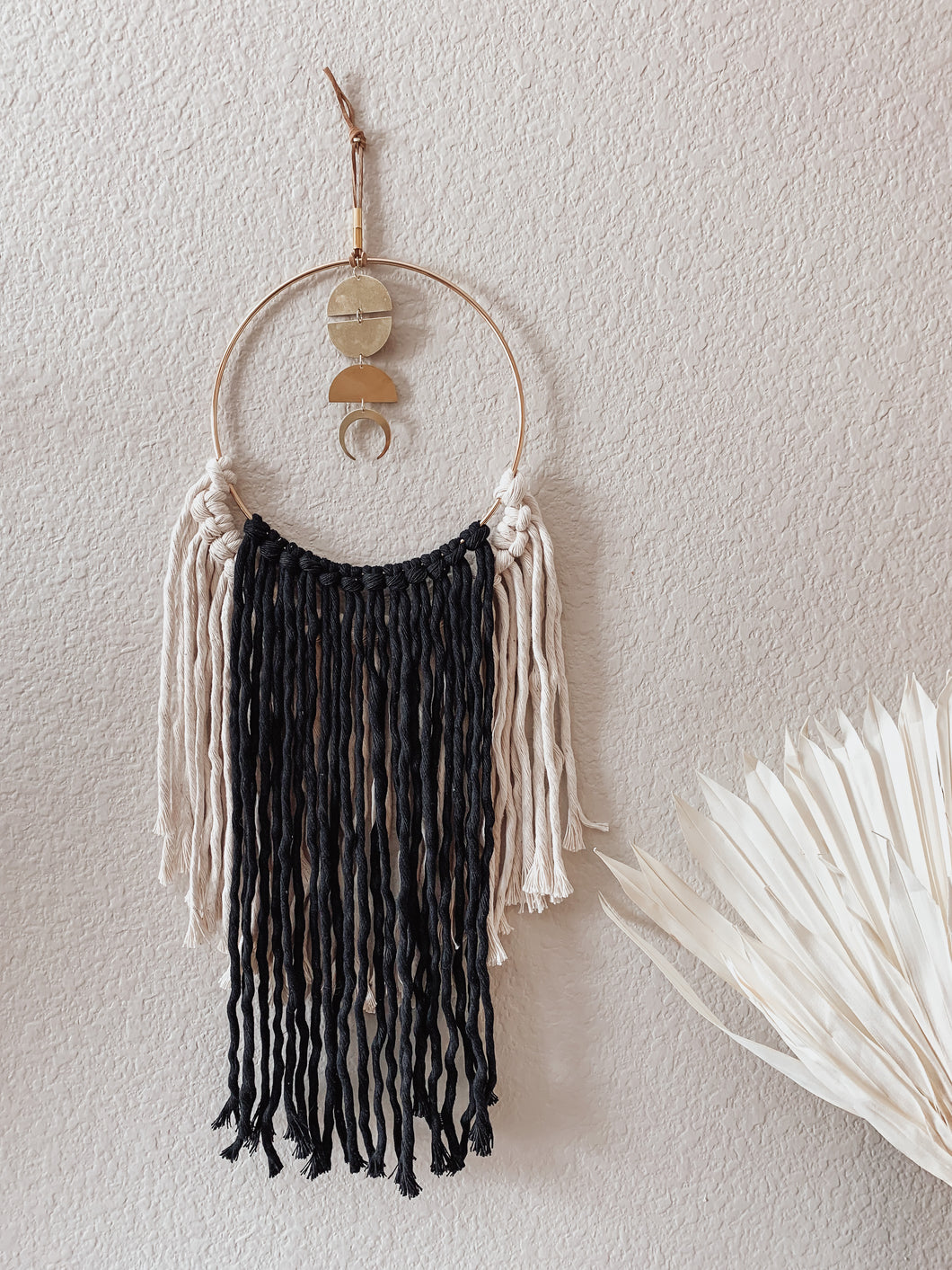 A macrame wall hanging with natural white and black colored fringe hand knotted on a gold ring with brass charms perfectly placed