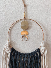Load image into Gallery viewer, A close up of a macrame wall hanging with natural white and black colored fringe hand knotted on a gold ring with brass charms perfectly placed
