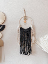 Load image into Gallery viewer, A macrame wall hanging with natural white and black colored fringe hand knotted on a gold ring with brass charms perfectly placed
