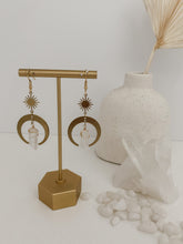 Load image into Gallery viewer, A pair of drop earrings with raw brass crescent and clear quartz charm seen on a jewelry stand
