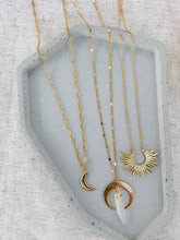 Load image into Gallery viewer, Flat lay of three layered 24K gold chain necklaces one with gold crescent charm, one with a gold crescent and clear quarts charm, and one with gold sun burst pendant.
