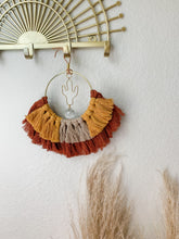 Load image into Gallery viewer, A macrame suncatcher with gold cactus charm and mix of maroon, mustard yellow, and tan cotton fringe hanging on a decorative wall hook
