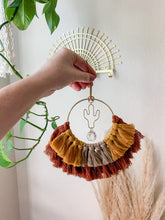 Load image into Gallery viewer, A hand holding a macrame suncatcher with gold cactus charm and mix of maroon, mustard yellow, and tan cotton fringe
