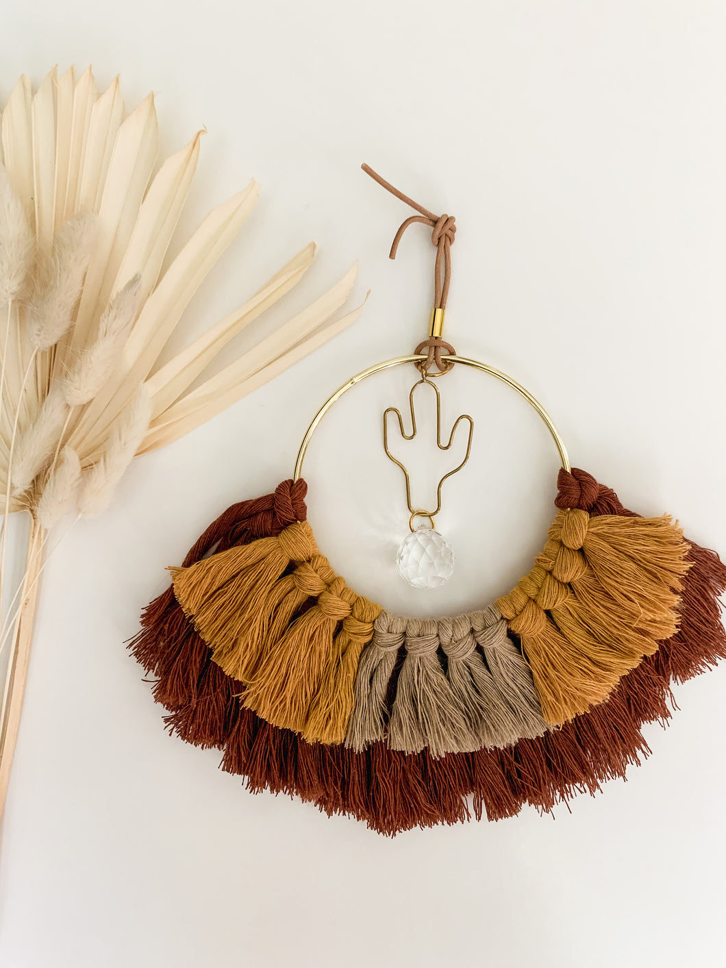 A macrame suncatcher with gold cactus charm and mix of maroon, mustard yellow, and tan cotton fringe