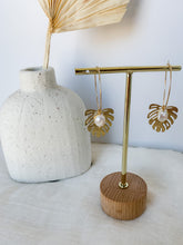 Load image into Gallery viewer, A pair of gold plated 30mm hoop earrings with gold plated monstera charm and freshwater pearl  on a jewelry stand
